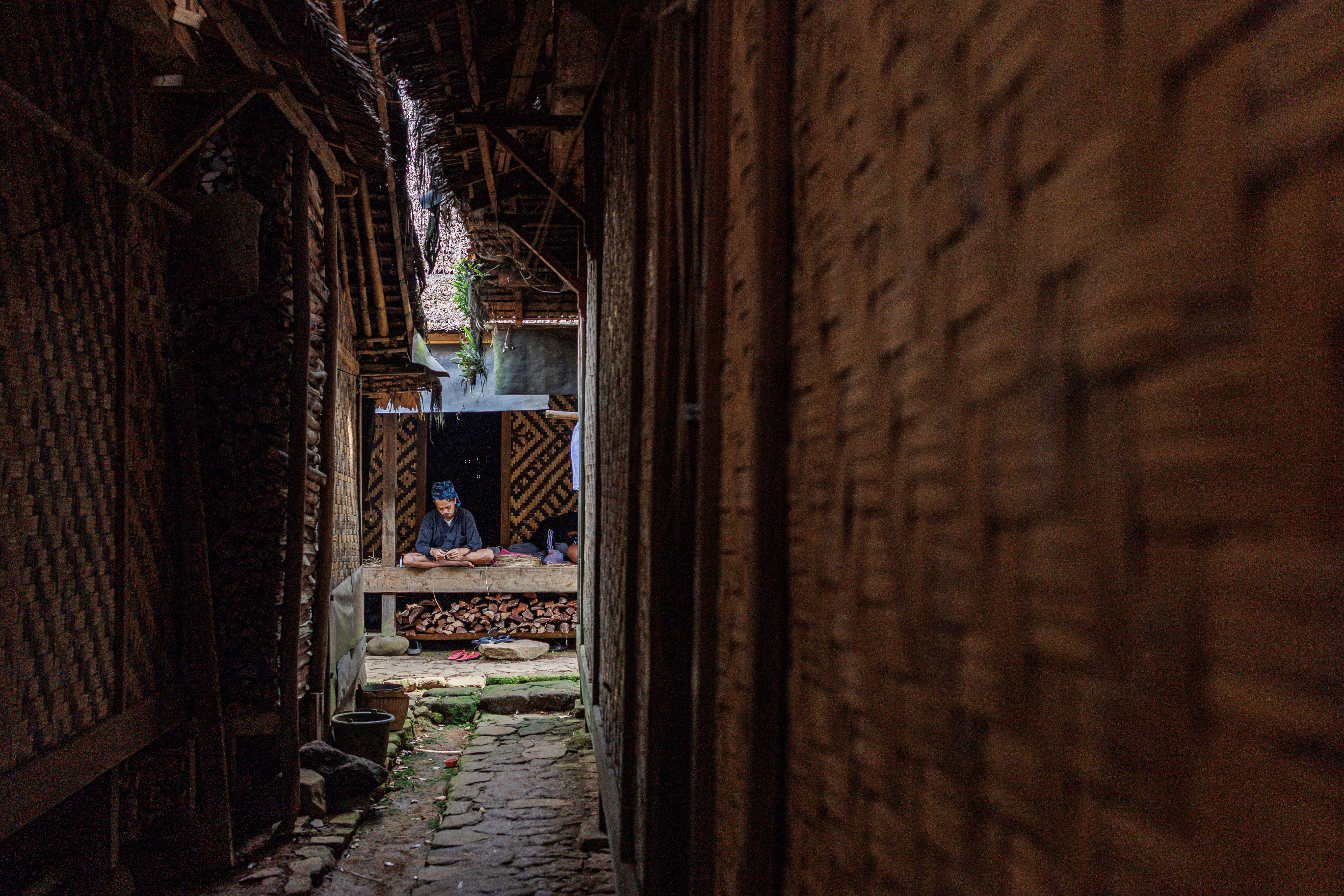 A man sits in a traditional Banten, Indonesian home's hallway, surrounded by woven bamboo walls.