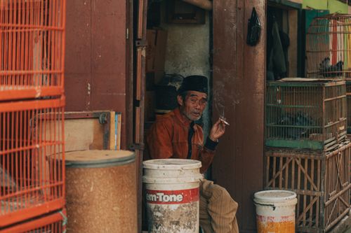 Free A local man in Jawa Barat, Indonesia, enjoys a cigarette outside a rustic shop surrounded by colorful bird cages. Stock Photo
