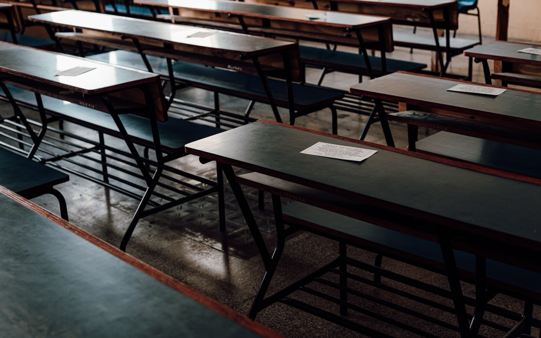A row of unoccupied desks and benches in a classroom with exam papers on top.