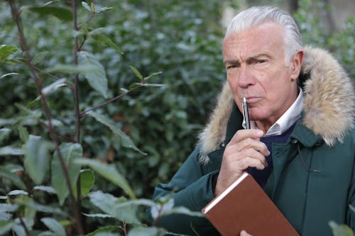 Man in Green Jacket Holding Brown Book and Pen