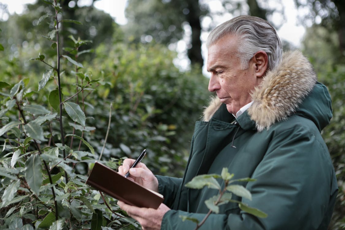 Side view of aged gray haired male in warm outerwear writing with pen in notebook while standing in park