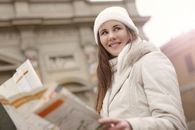 From below side view of cheerful female tourist in warm clothes standing on street and reading paper map during sightseeing trip in city