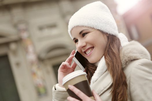 一位穿着保暖衣服的女性侧身站在街上，端着热饮，一边微笑一边用手机聊天，目光移向别处