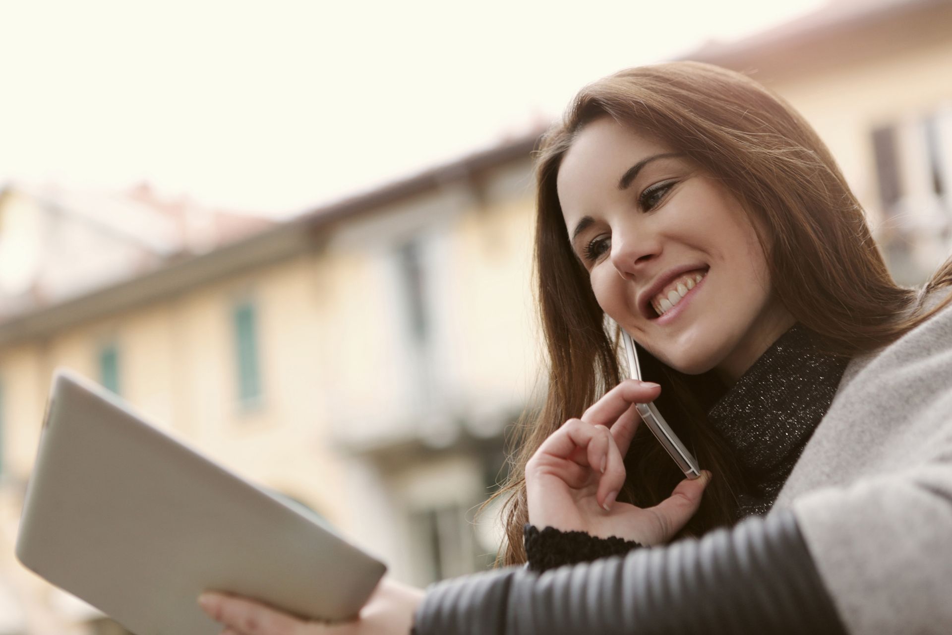 Smiling woman talking on a smartphone and holding a tablet outdoors, showcasing modern technology use.