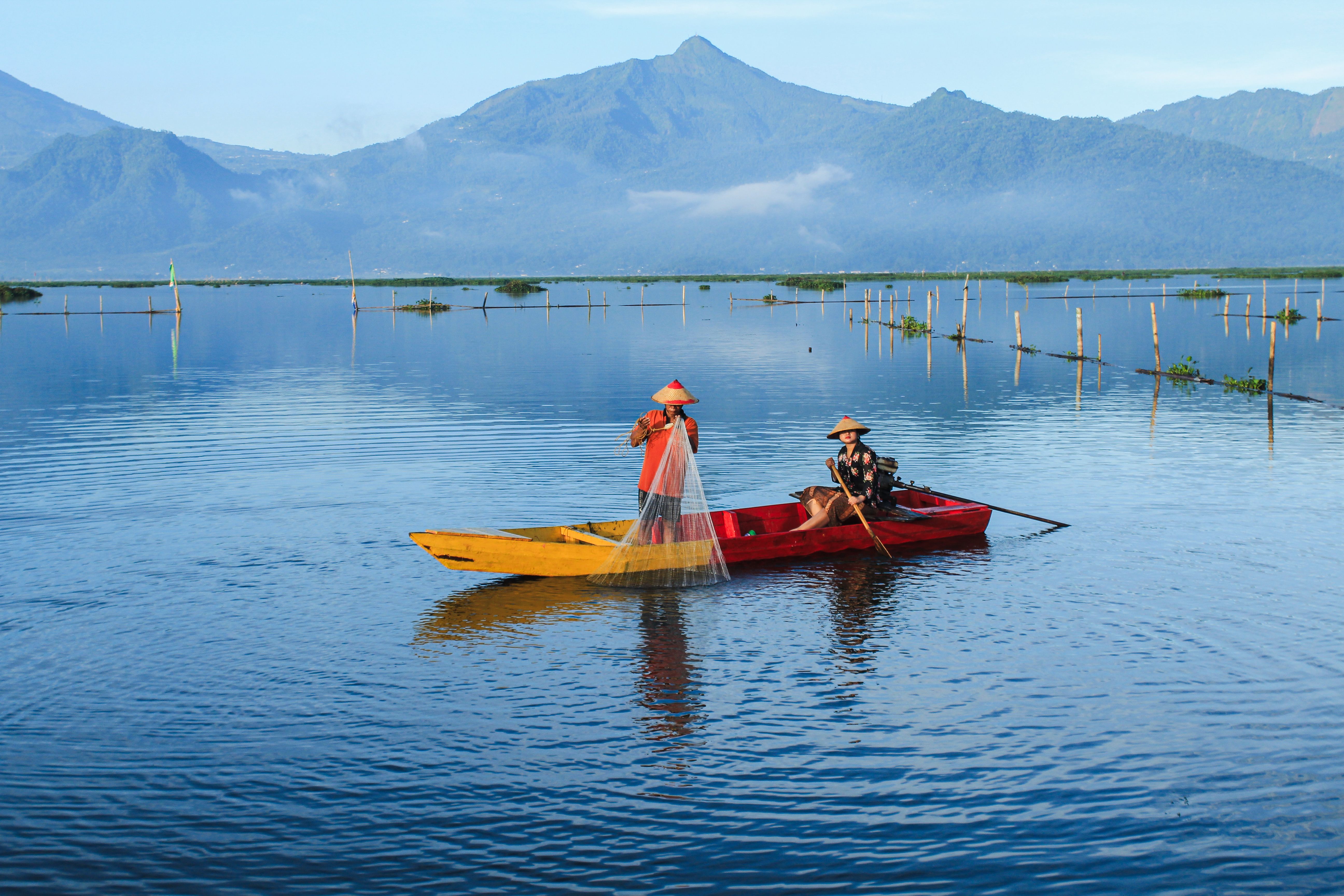 Traditional fishermen in a canoe casting nets at Rawa Pening, Central Java, Indonesia, against a mountain backdrop.