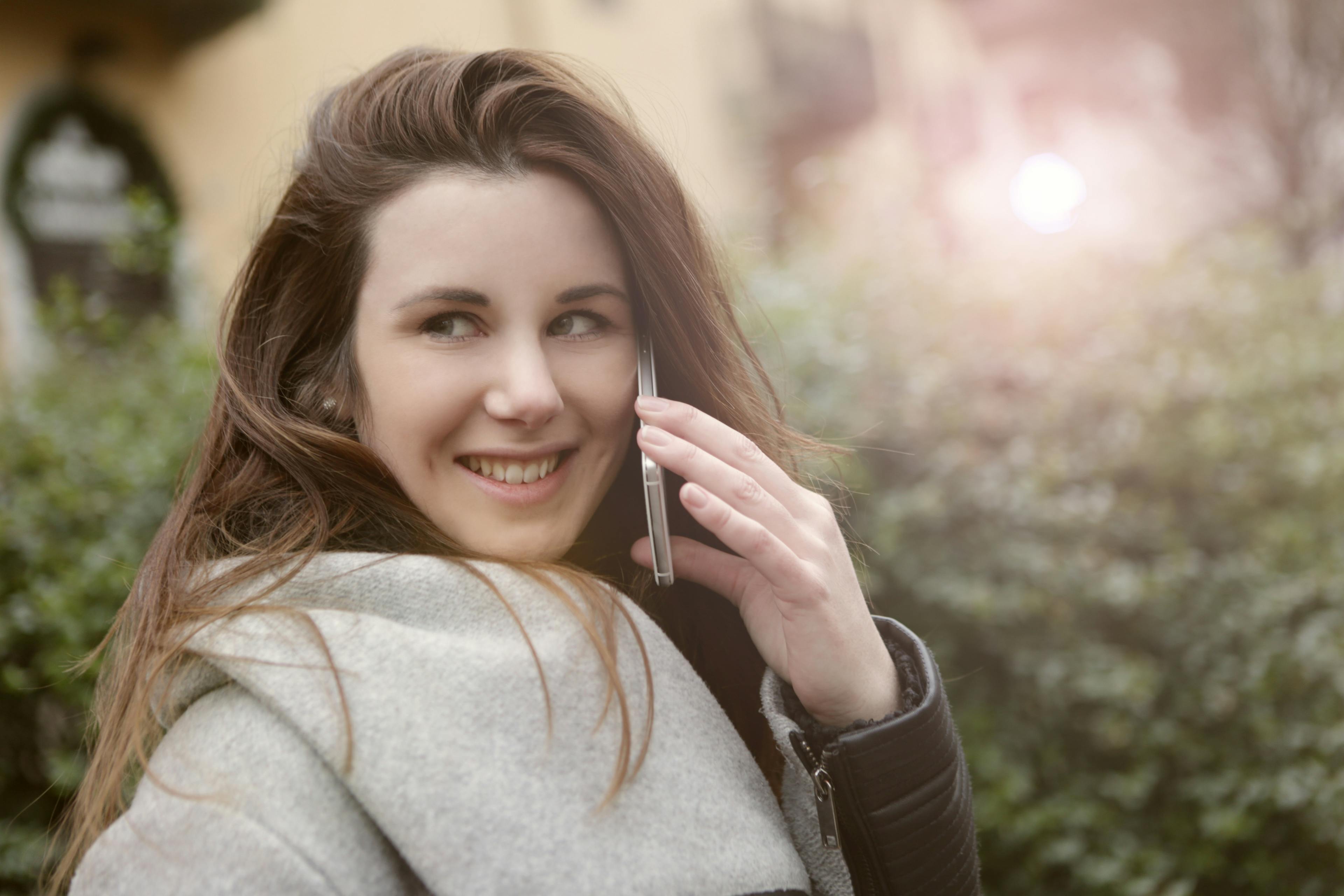 A relaxed young woman smiling while talking on her smartphone in a sunny outdoor setting.