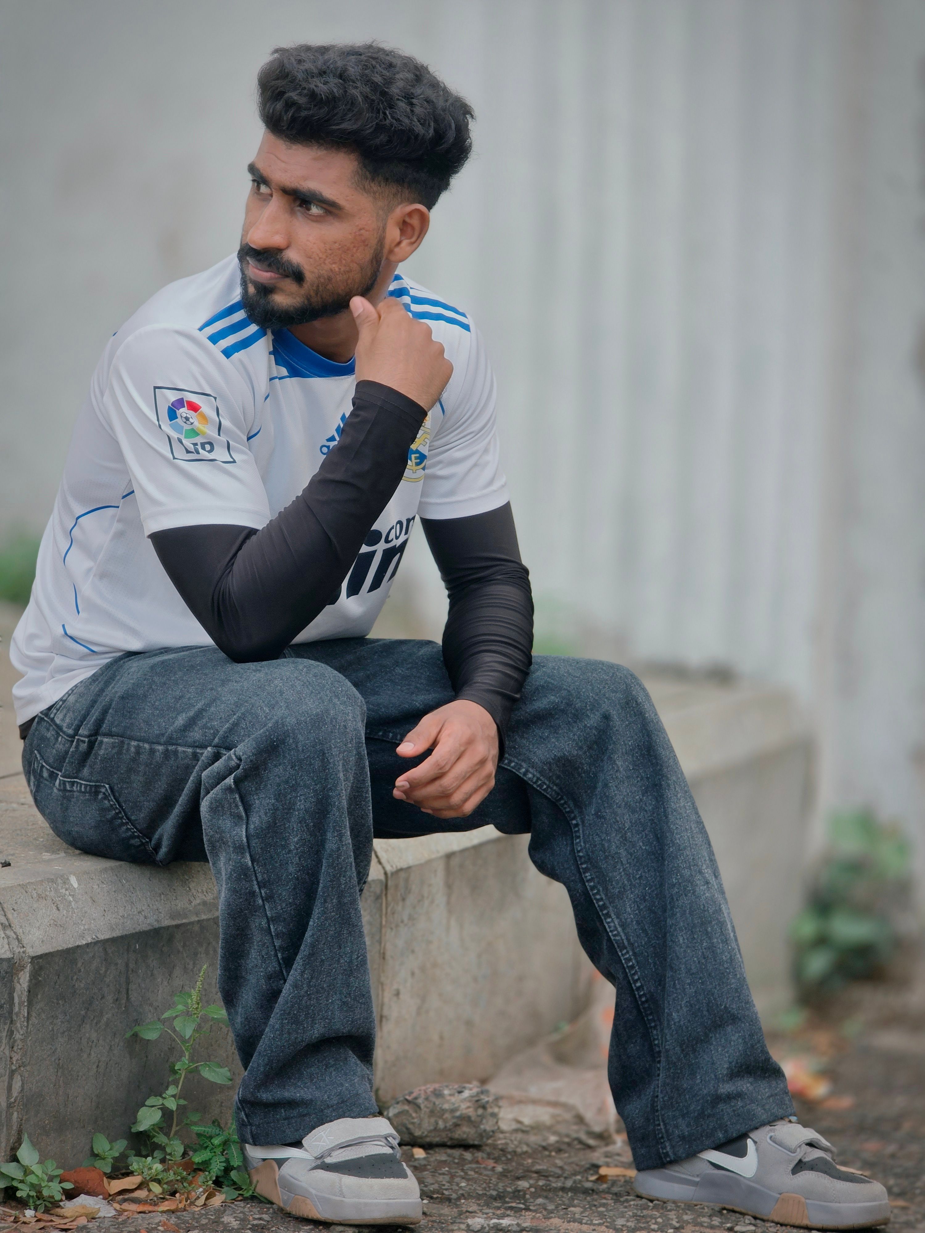 A thoughtful man sits outdoors on a concrete ledge, wearing a casual outfit.