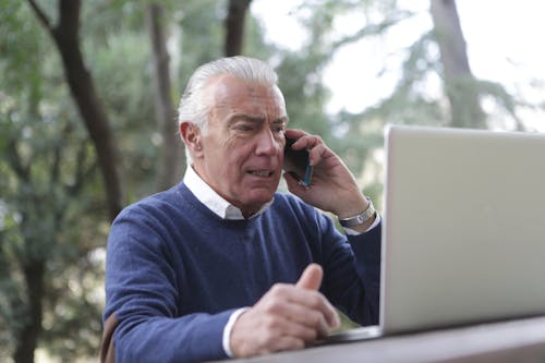 Man in Blue Sweater Sitting by the Table Using Laptop and Cellphone