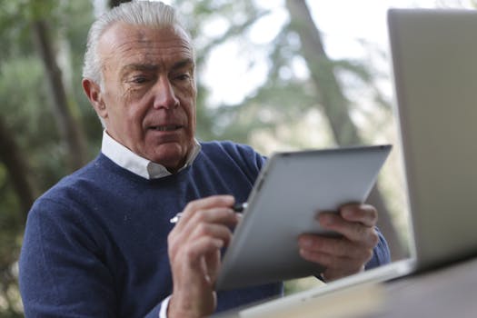 Elderly man focused on using a tablet in a natural outdoor setting.