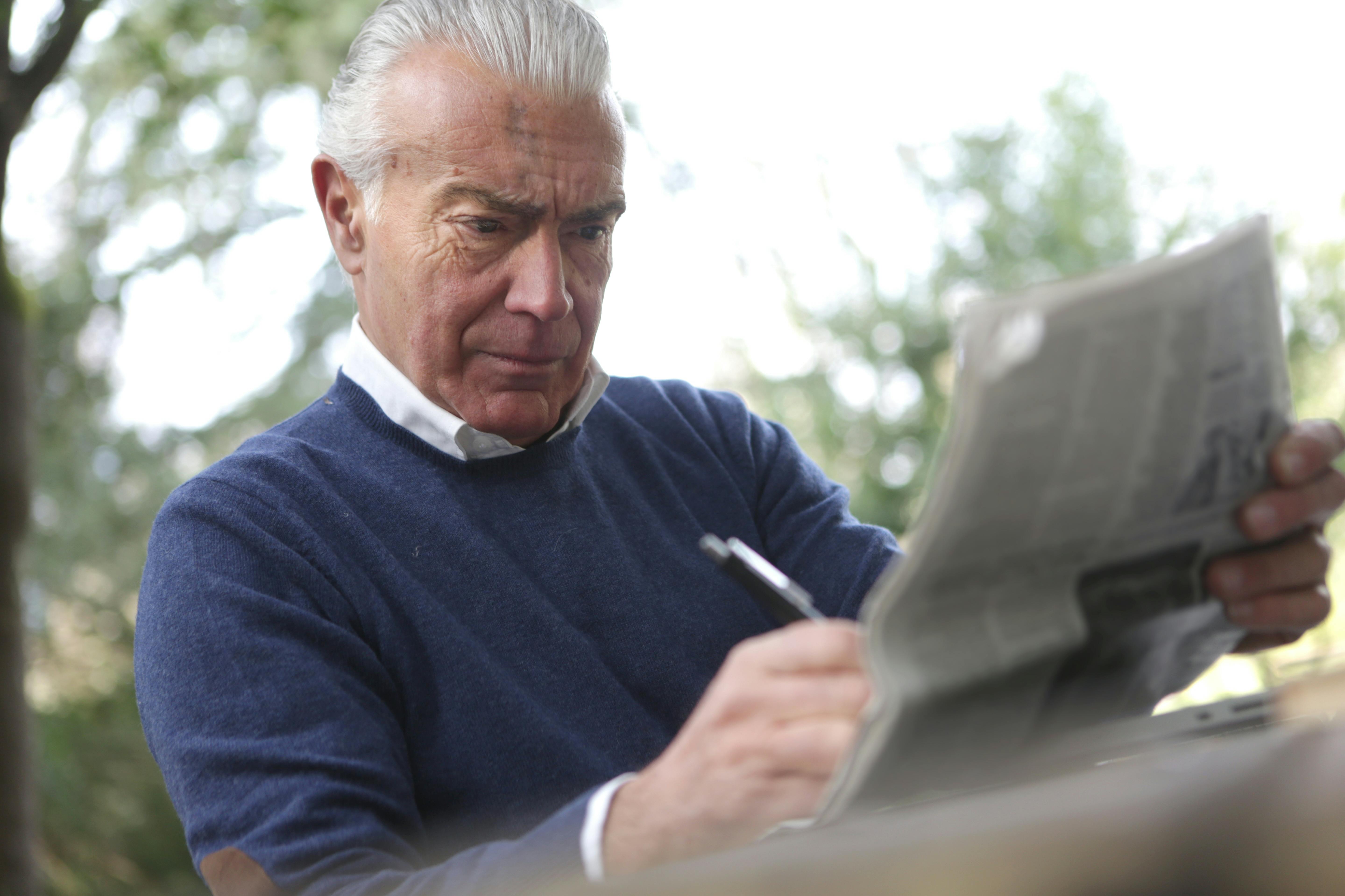 man in blue sweater holding pen and reading newspaper