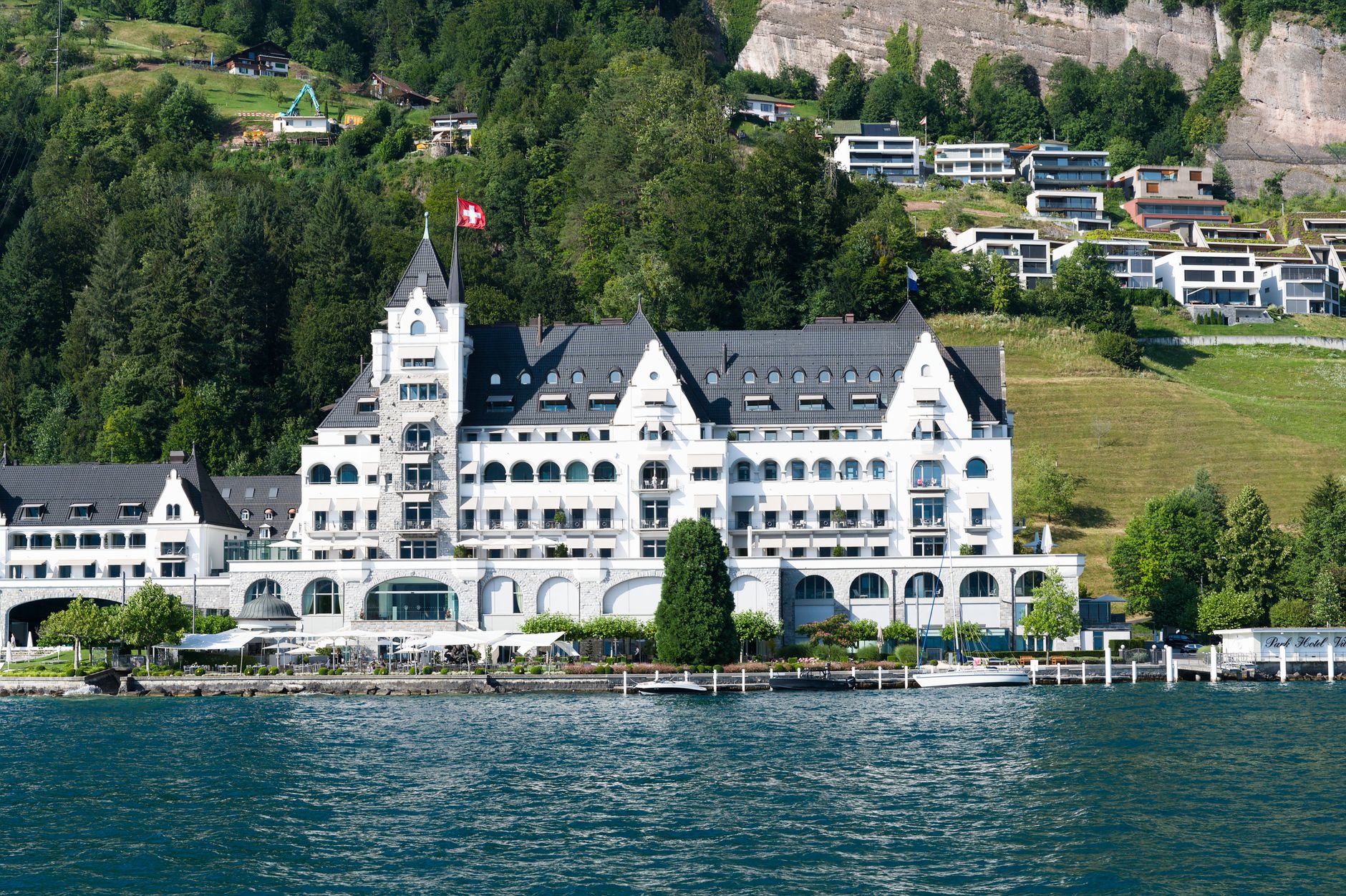 Stunning Swiss hotel by the lakeside with lush mountains and a clear blue sky.
