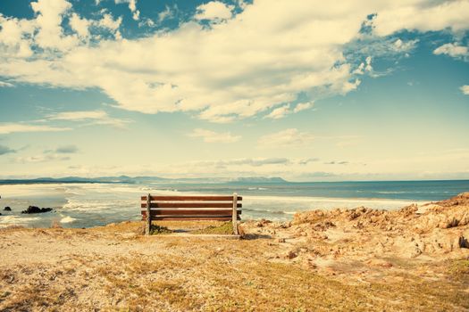 Solitary bench overlooking a serene ocean with distant mountains under a blue sky with clouds.