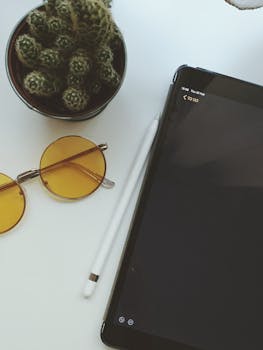 Flatlay of a trendy workspace with a cactus, tablet, stylus, and sunglasses on a white table.