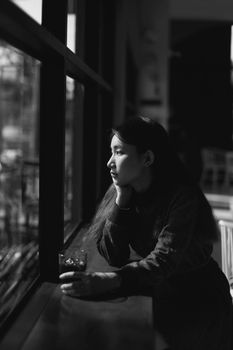Black and white portrait of a woman thoughtfully looking outside a cafe window.