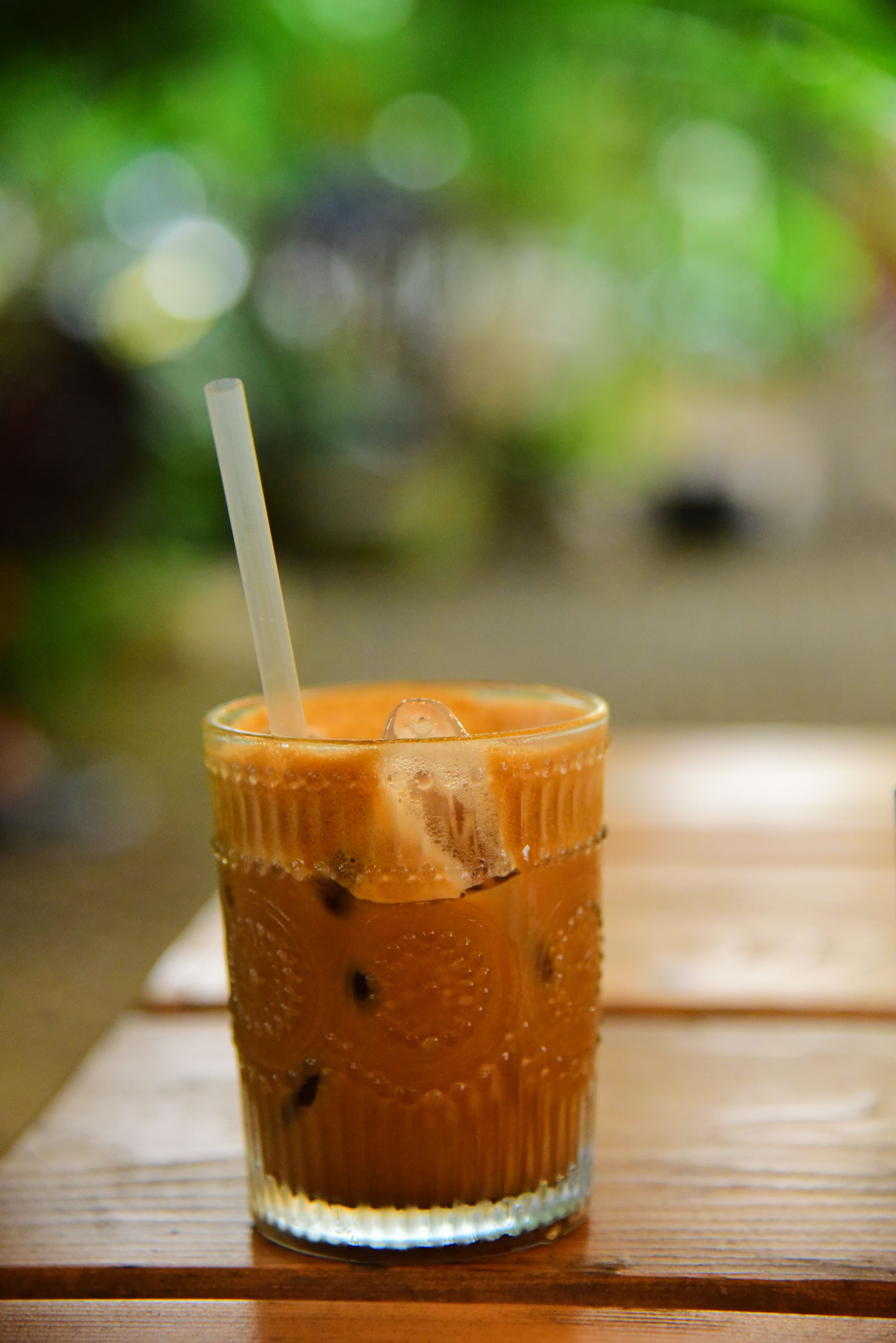 A glass of iced coffee with straw on a wooden table outdoors, surrounded by nature.