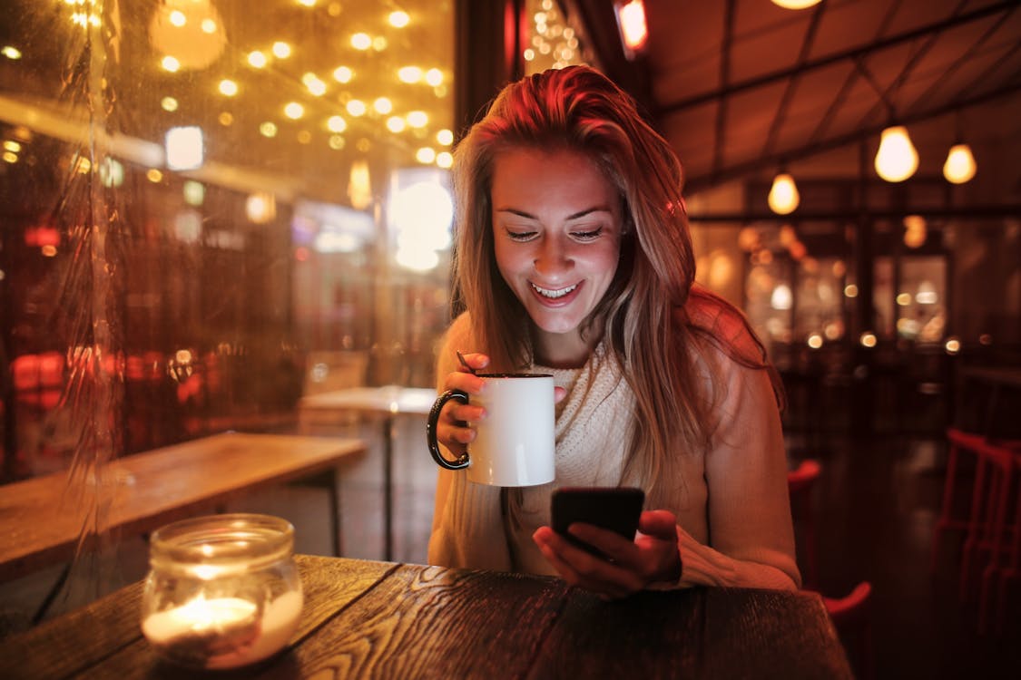 Free Woman Holding White Ceramic Mug Looking at Her Cellphone Stock Photo
