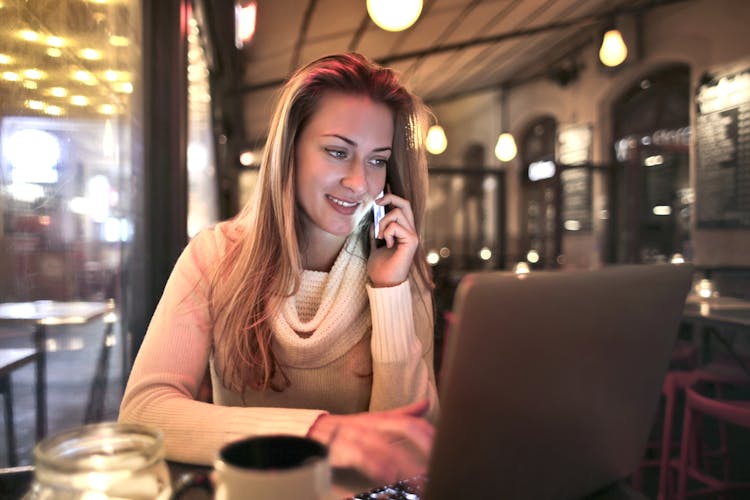 Relaxed Woman In Cafe Talking On Smartphone And Using Laptop