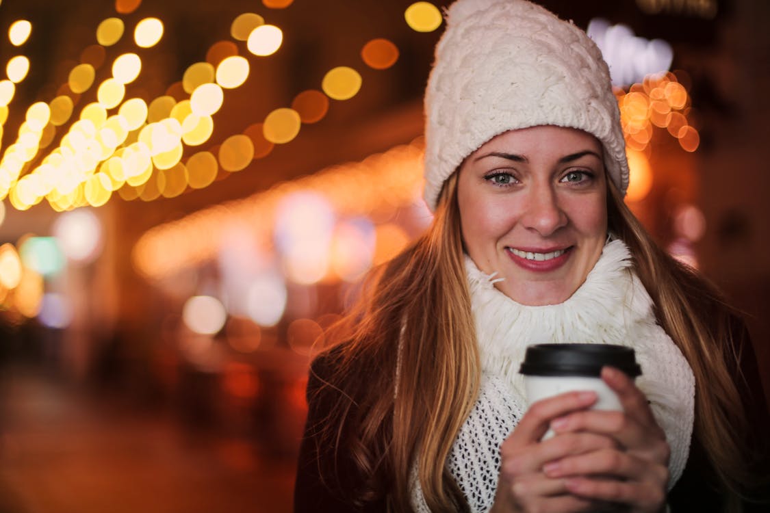 Free Cheerful female in warm outerwear standing with beverage to go on street and chilling during weekend in evening in city illumination while looking at camera Stock Photo