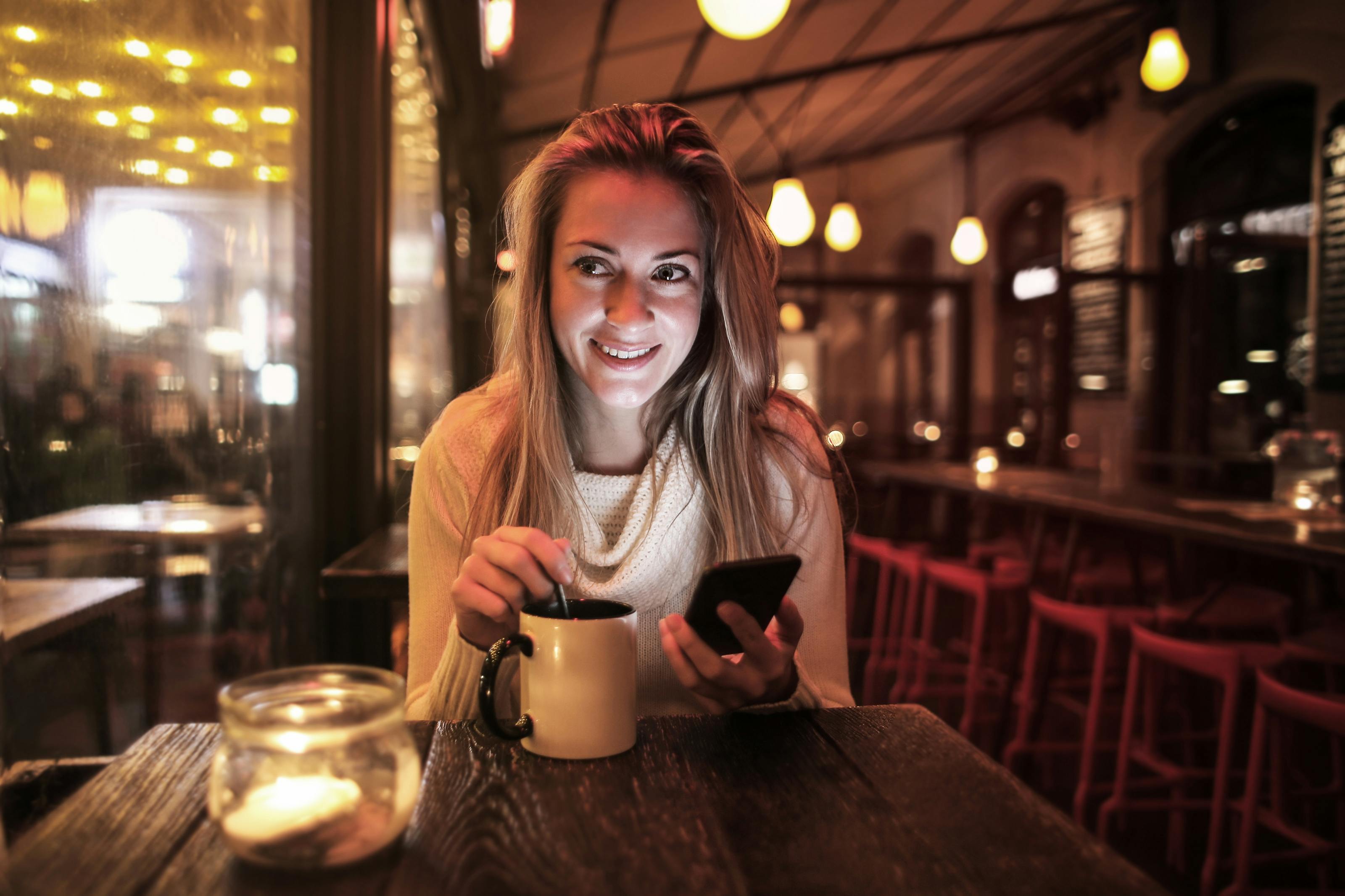 delighted woman with cup of beverage browsing smartphone in cafe