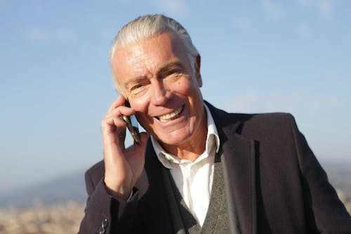 Free Delighted male entrepreneur wearing classy jacket standing in city and making phone call while smiling and looking at camera Stock Photo