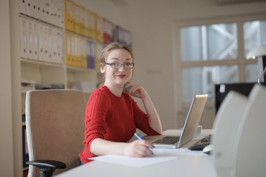 Smiling woman in glasses working on a laptop at a desk in a bright, modern office.