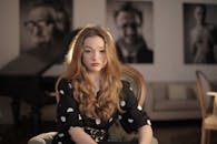 一位留着长波浪卷发的女子坐在室内，在模糊的背景前露出沉思的表情