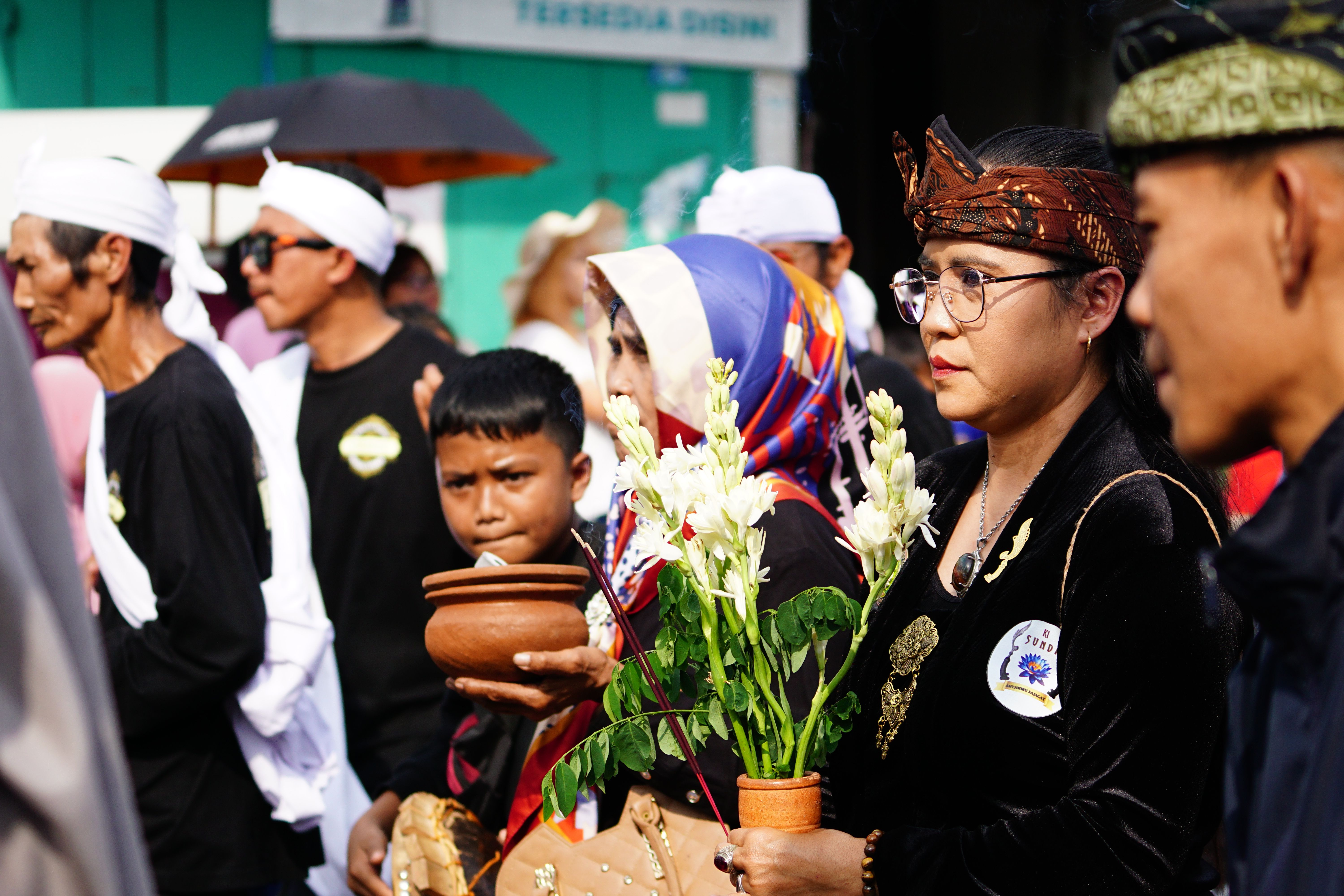 A group participates in a cultural procession wearing traditional attire, holding flowers and pottery.