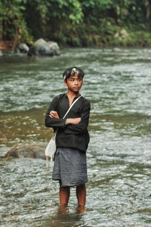 Free A young boy stands in a river in Banten, Indonesia, wearing traditional attire and carrying a bag. Stock Photo