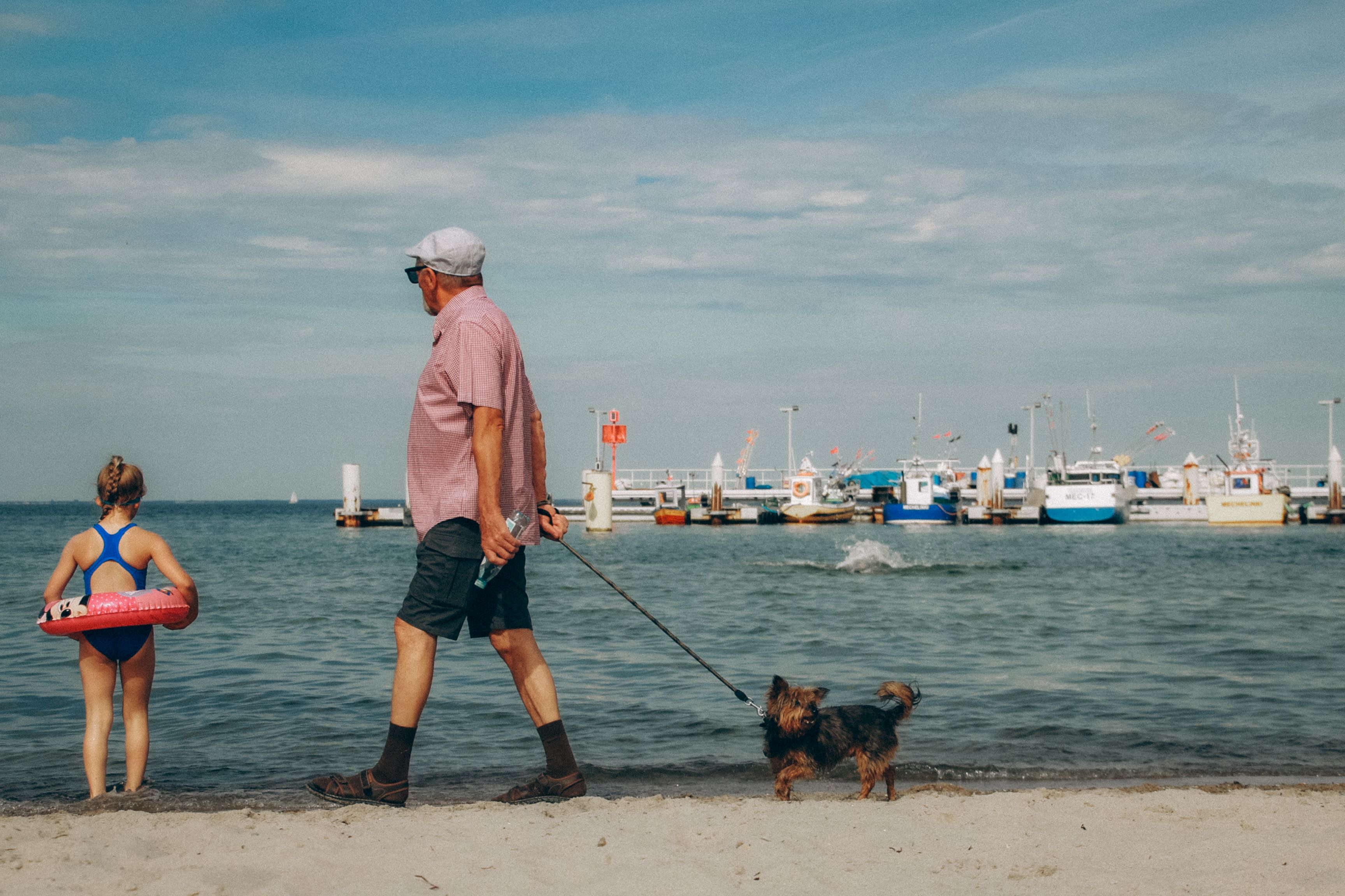 A senior man walks a dog while a child plays near the water at a beach.