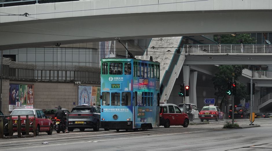 Iconic Hong Kong tram traverses a wet urban street under a bridge on a rainy day.