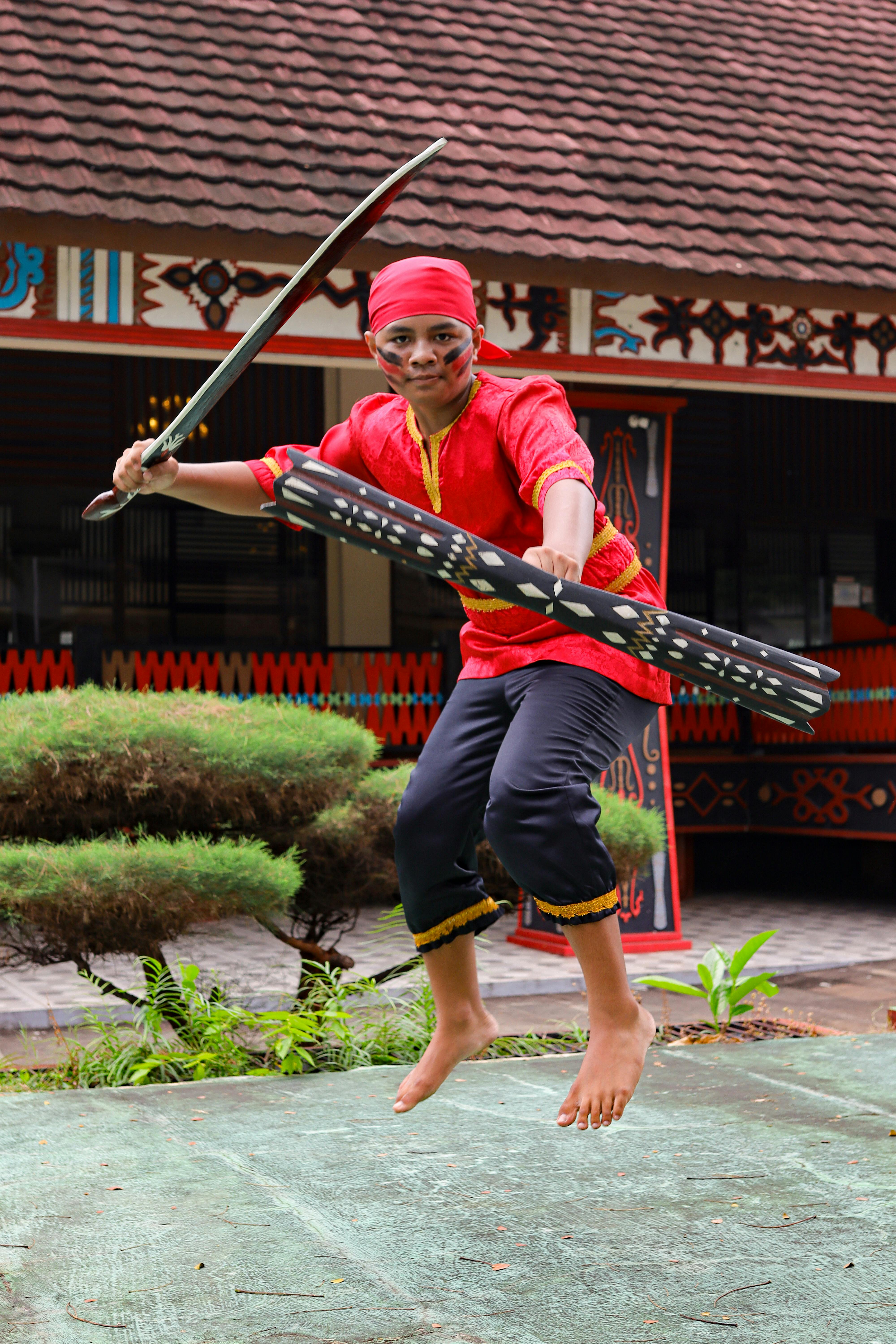 A young man in traditional Indonesian costume leaps while holding a sword and shield in a cultural dance performance.