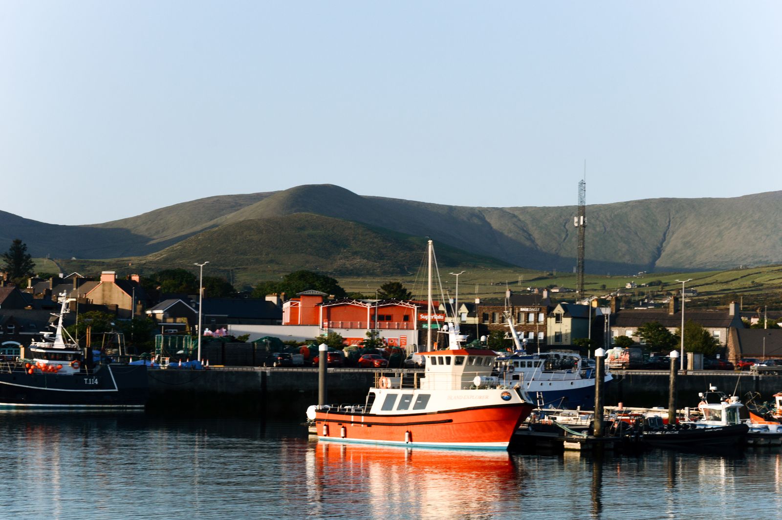 A view of Dingle Harbor in Ireland featuring fishing boats and green hills under a clear sky