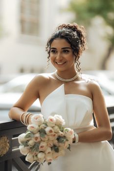 A smiling bride holding a bouquet of roses, posed outdoors in a radiant wedding dress.