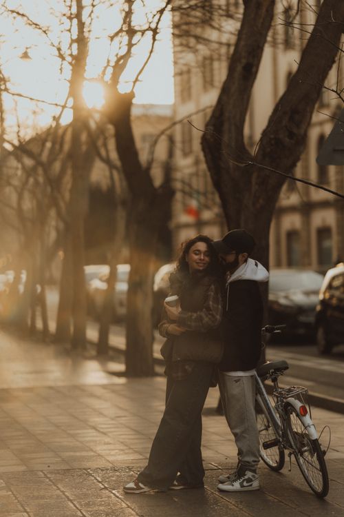 Free A couple embraces on a tree-lined city street during a warm sunset, with a bicycle nearby. Stock Photo