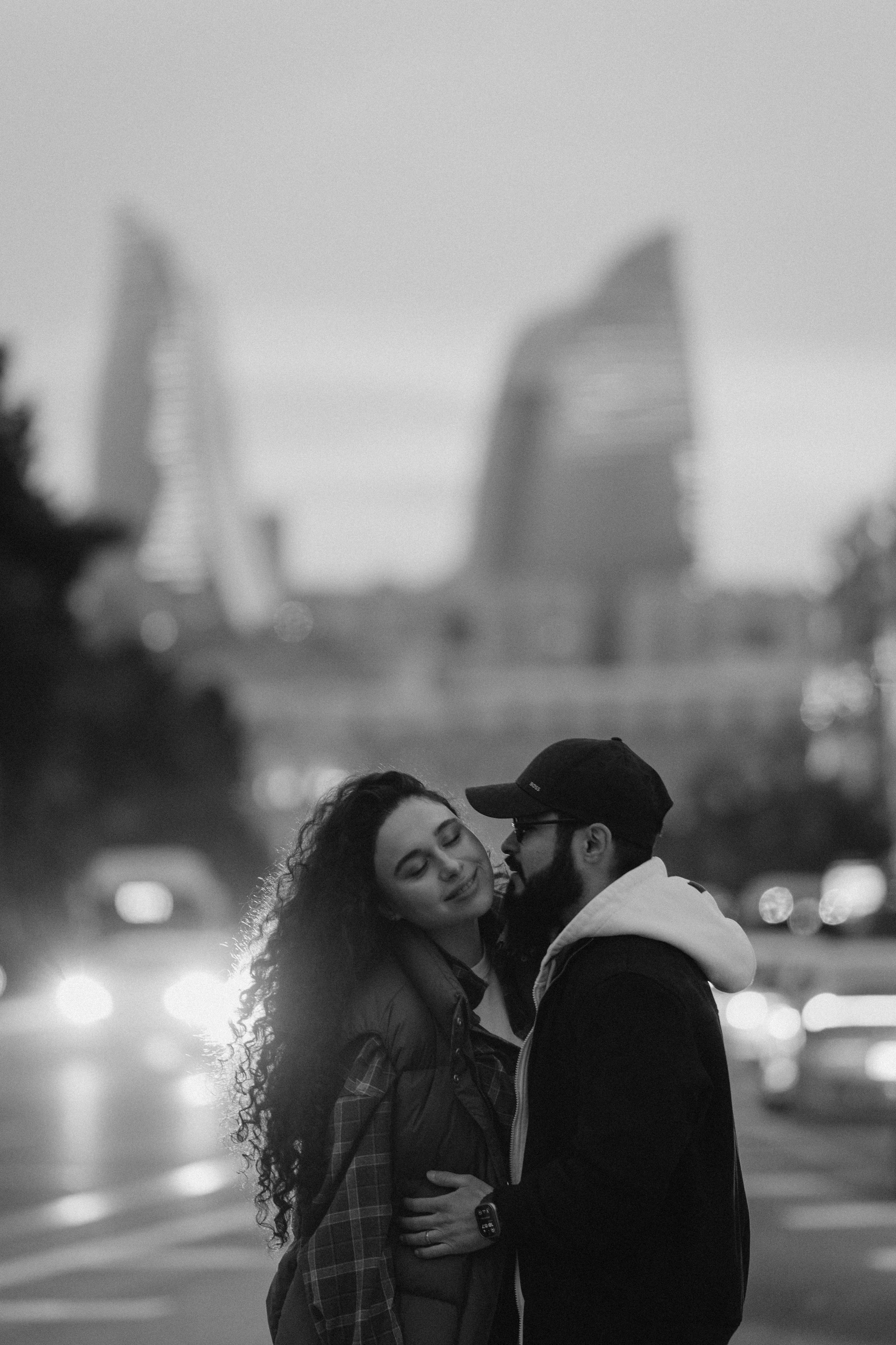 Black and white photo of a couple embracing in front of illuminated modern architecture.