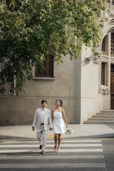 A happy couple in wedding attire walking across a city street crosswalk on a sunny day.