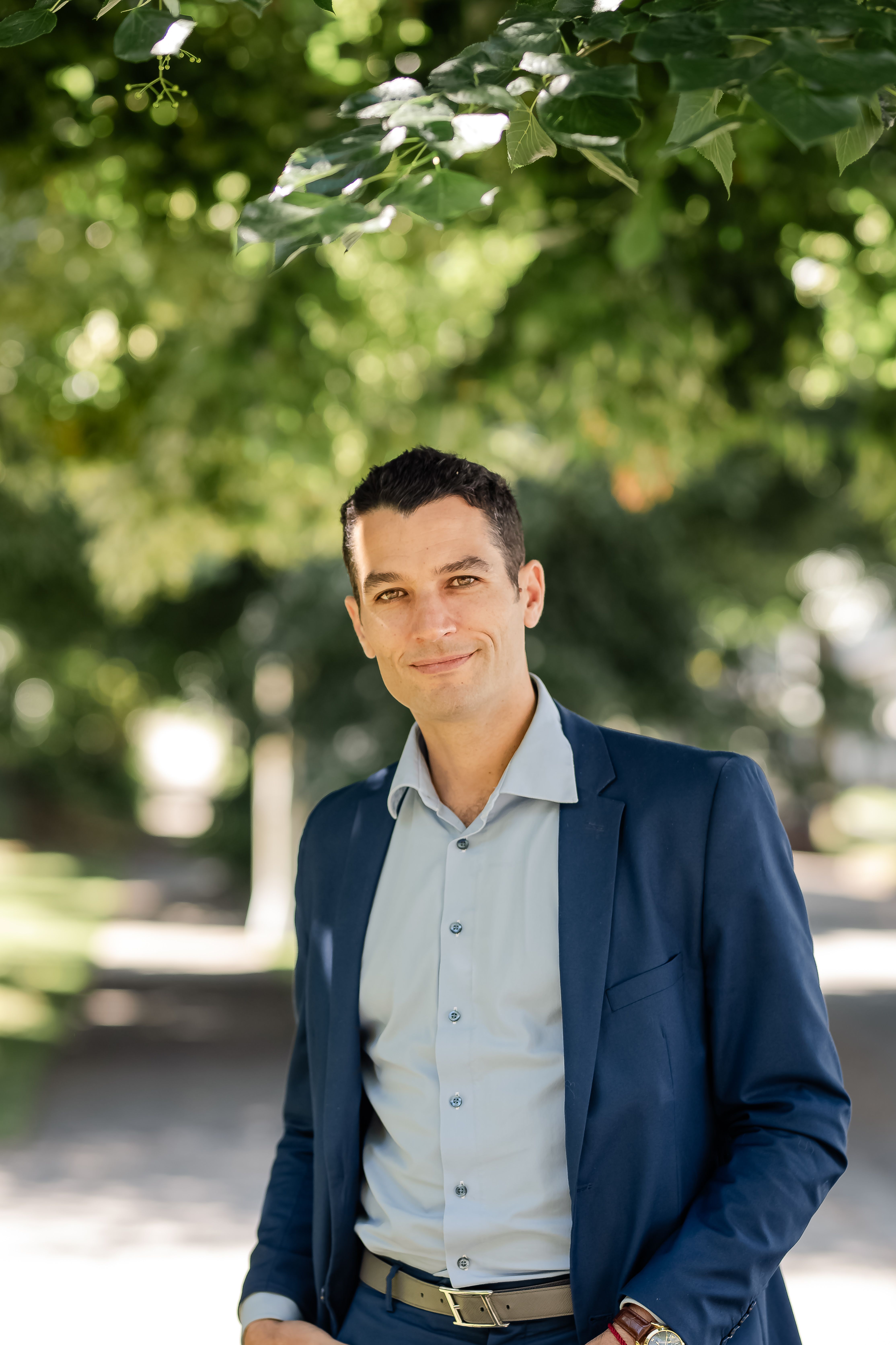 Professional businessman in a blue suit smiling confidently outdoors under green trees. Bright and friendly atmosphere.