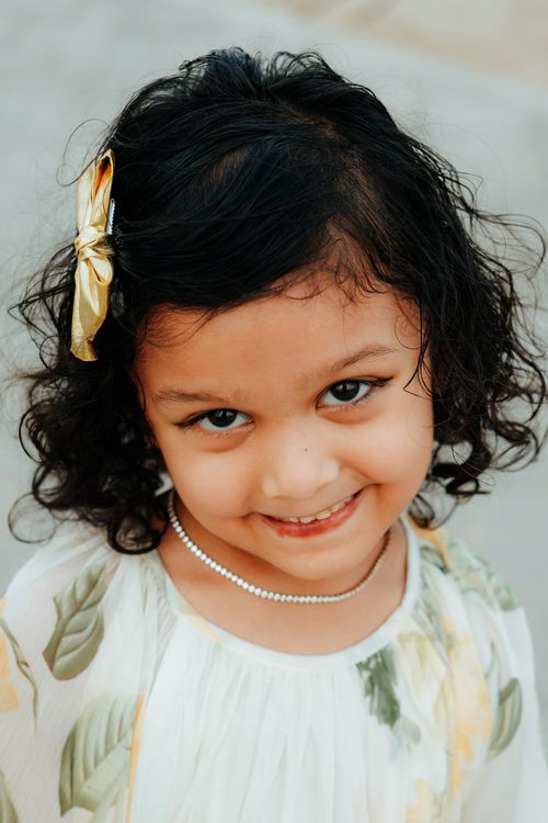 Free Adorable toddler with curly hair and bow smiling warmly in a floral dress, showcasing innocence and joy. Stock Photo