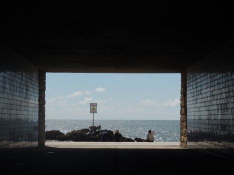 View from a tunnel leading out to a sunny ocean horizon with distant figures and warning sign.