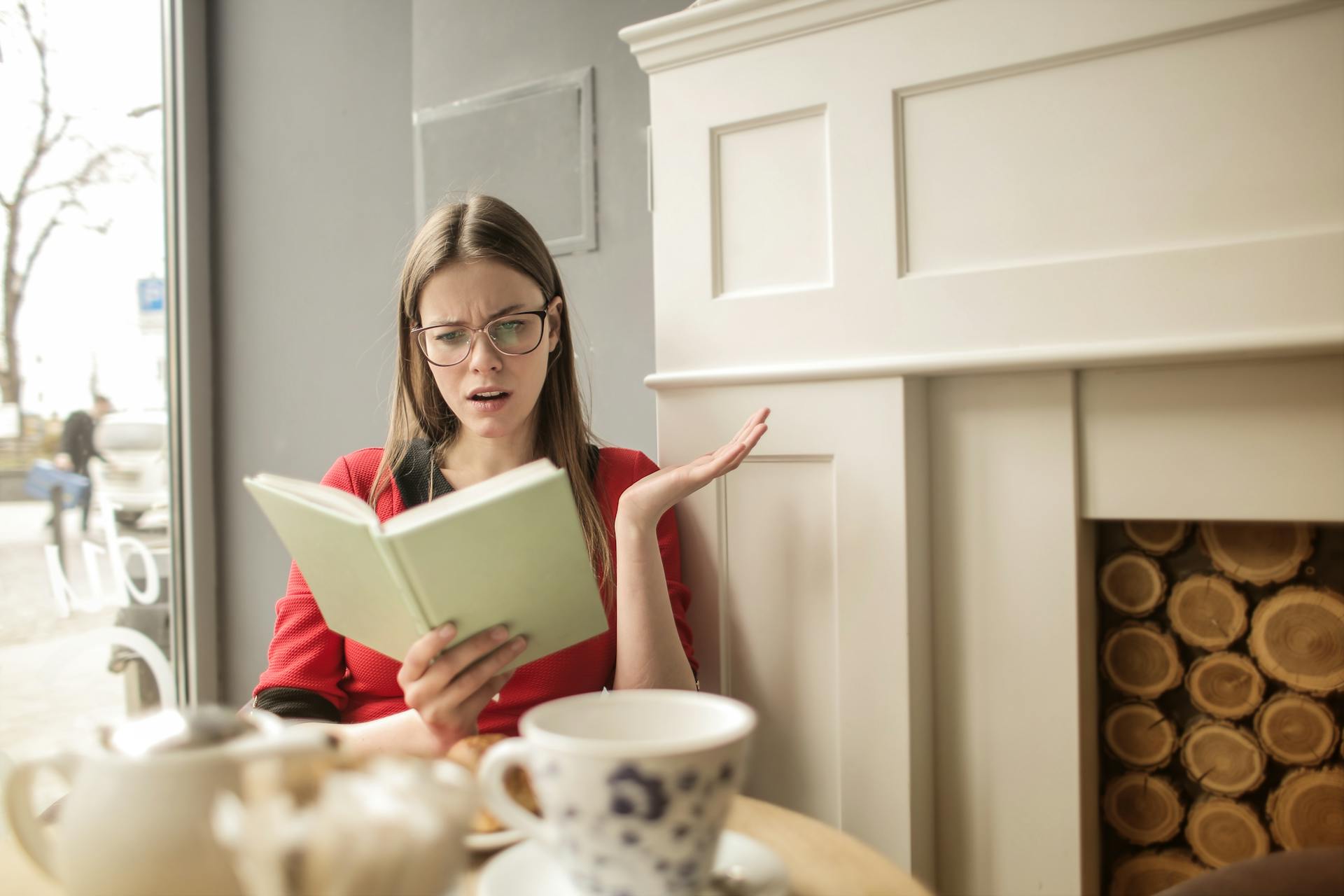 A woman reading and reacting to a book in a cozy cafe, surrounded by coffee and teapots.