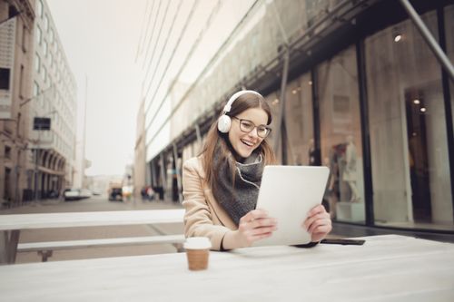 Free Cheerful woman with headphones using tablet at outdoor café in urban setting. Stock Photo