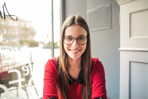 Free A smiling woman in eyeglasses and a red top, seated indoors by a window. Stock Photo