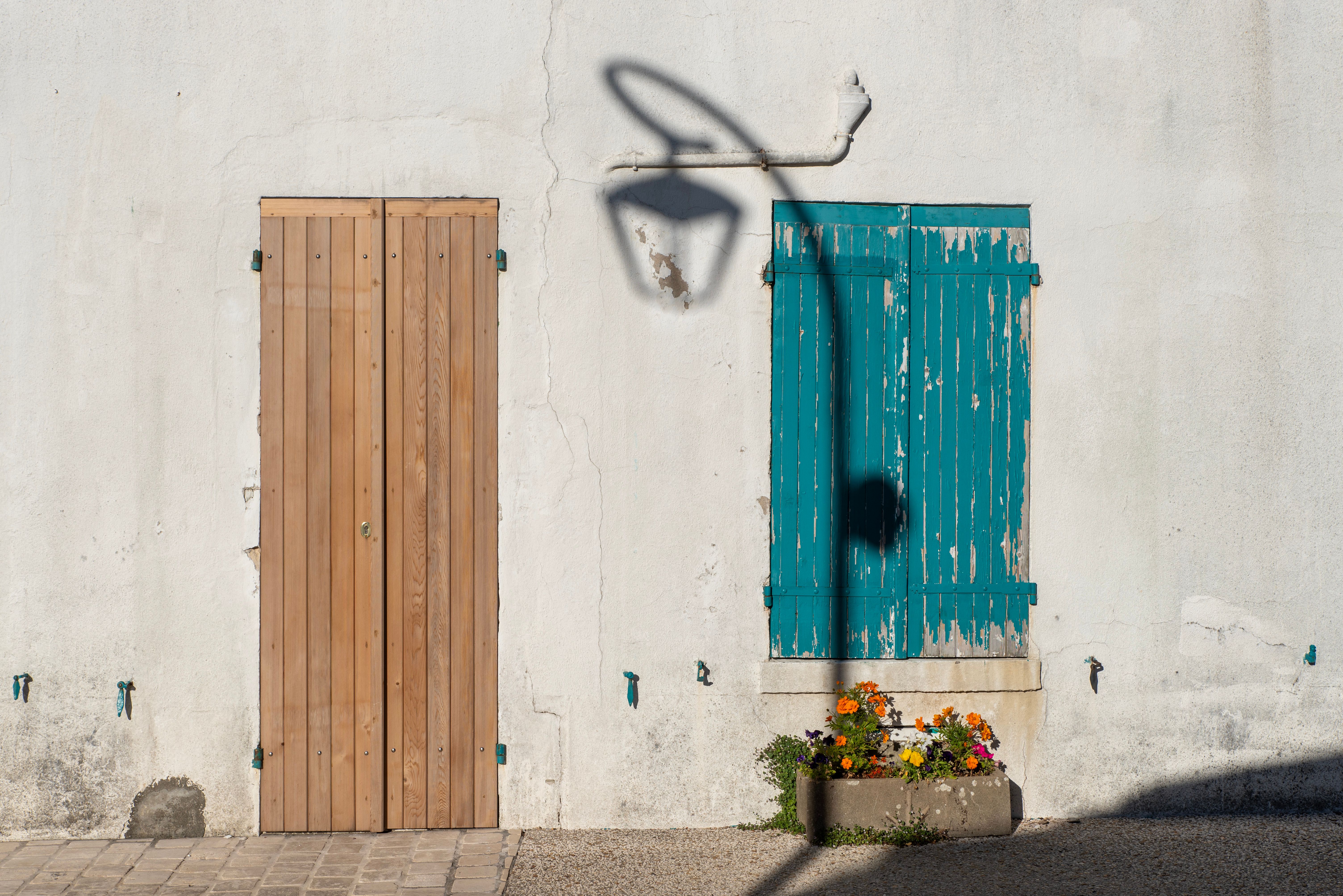 A rustic wooden door beside a blue shuttered window and street lamp shadow on a sunny day.