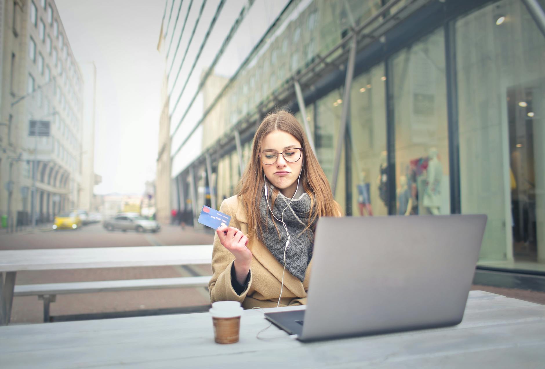 woman sat in coat and scarf out side with laptop and coffee with card in hand and looking stressed with her banking