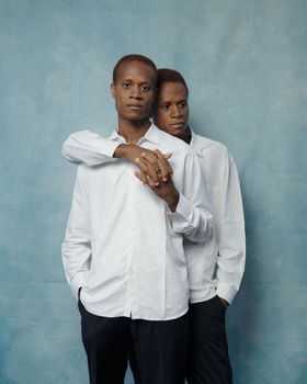 Two men in matching white shirts pose against a soft blue textured background, conveying unity.