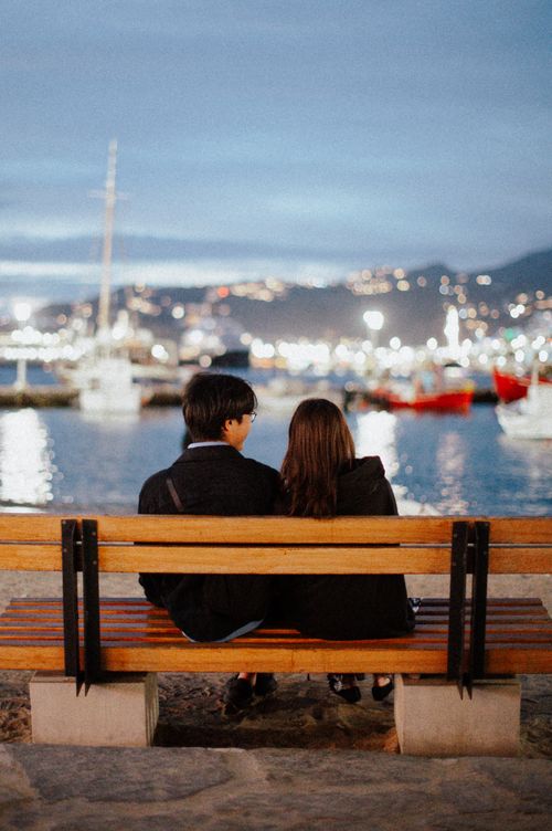 Free Couple enjoys a serene evening on a bench by the sea in Greece with city lights in view. Stock Photo