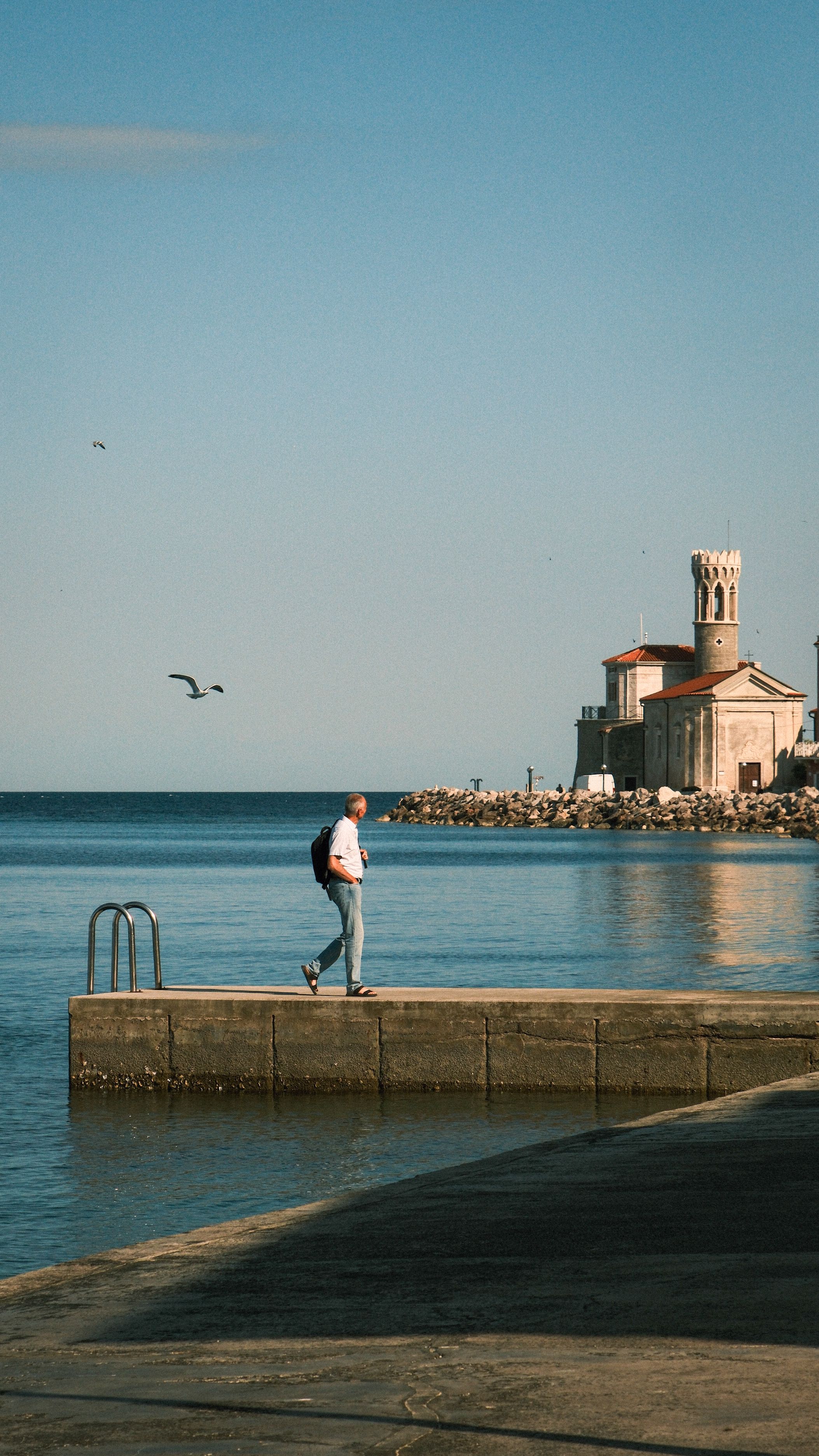 Man walking along a pier near a scenic seaside church under a clear sky.