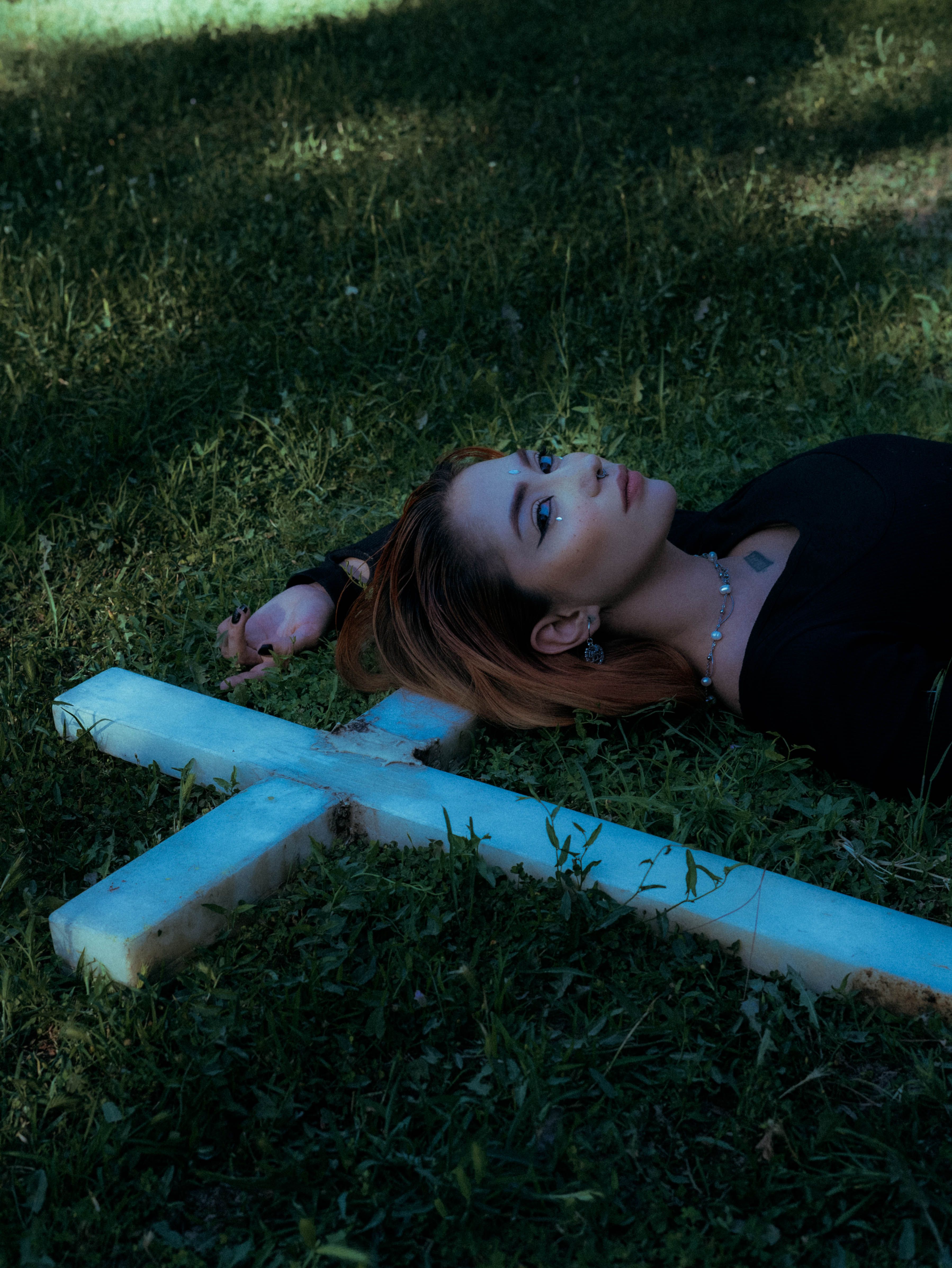 Young woman lying on grass beside a stone cross, evoking contemplation and peace.