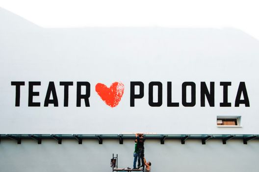 Workers on a lift painting the 'Teatr Polonia' sign on a large building exterior.