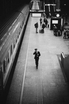 A train conductor walks through a bustling station in this classic black and white photograph.
